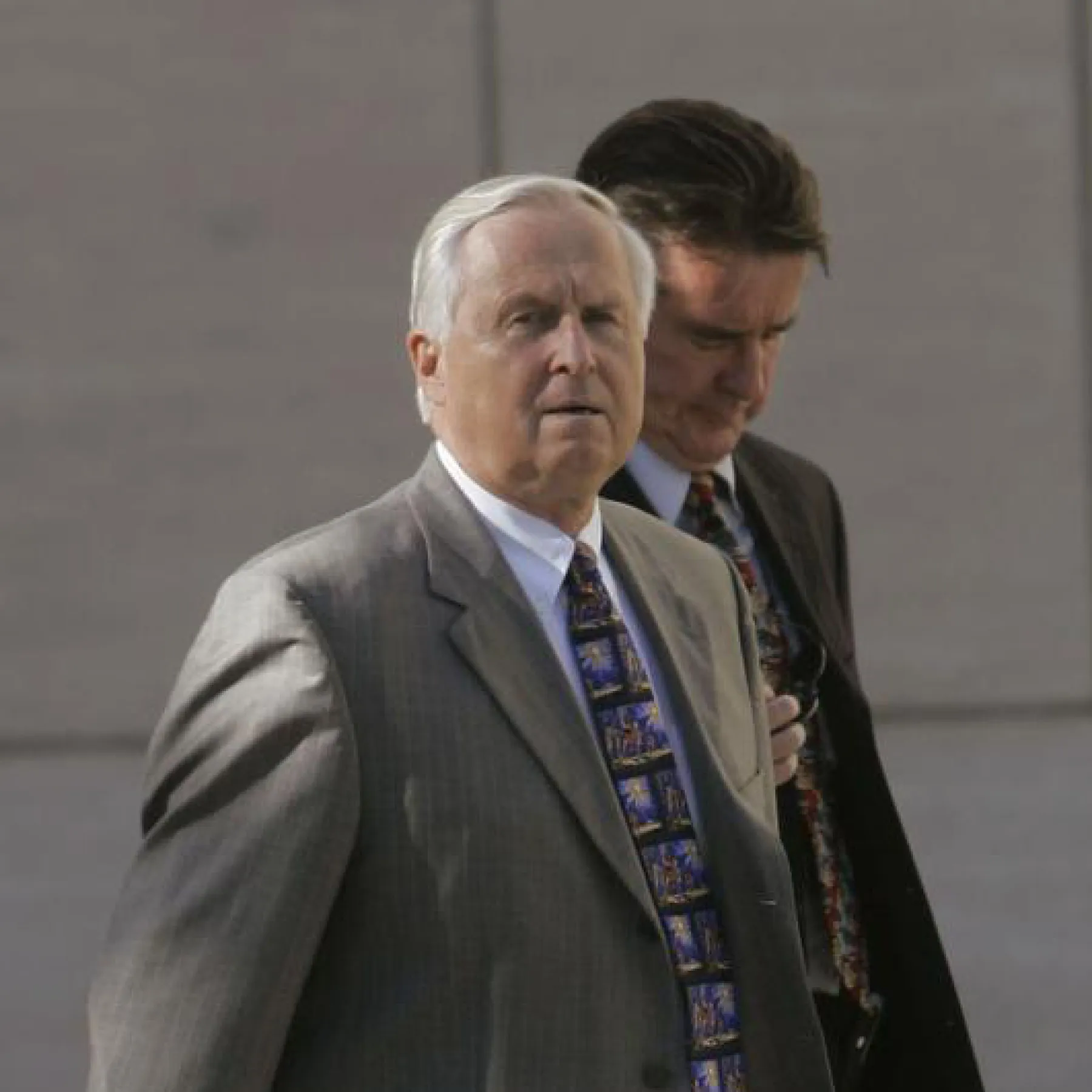 Two men in suits walk outdoors. The man in front has gray hair and wears a patterned tie, while the man behind him has brown hair and also wears a tie. They appear serious and focused, with a plain background behind them.
