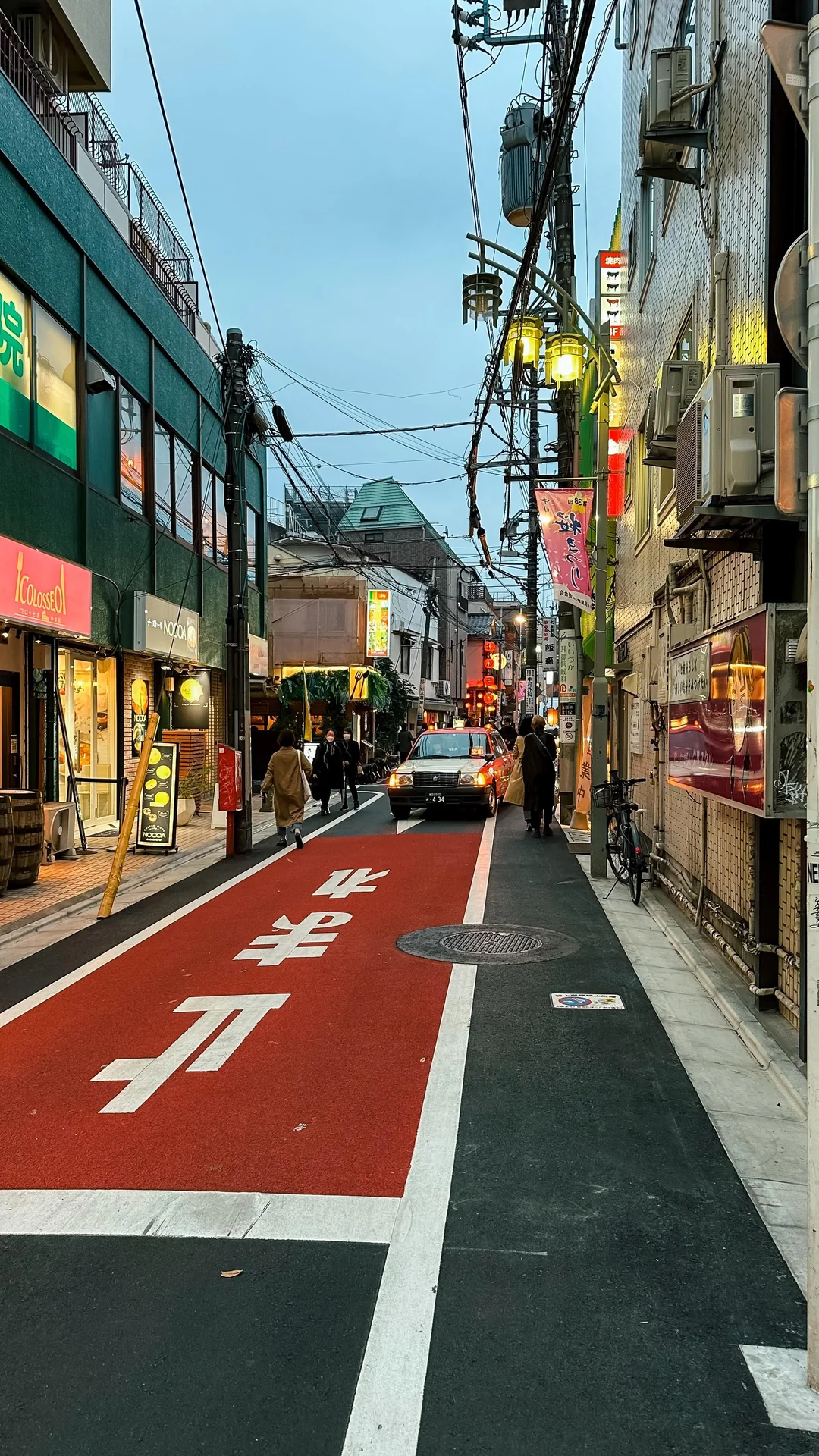 A narrow city street in Japan with red pavement and white Japanese characters, lined with shops, signs, and lights. People walk along the sidewalk, and a car is parked ahead under an overcast sky.