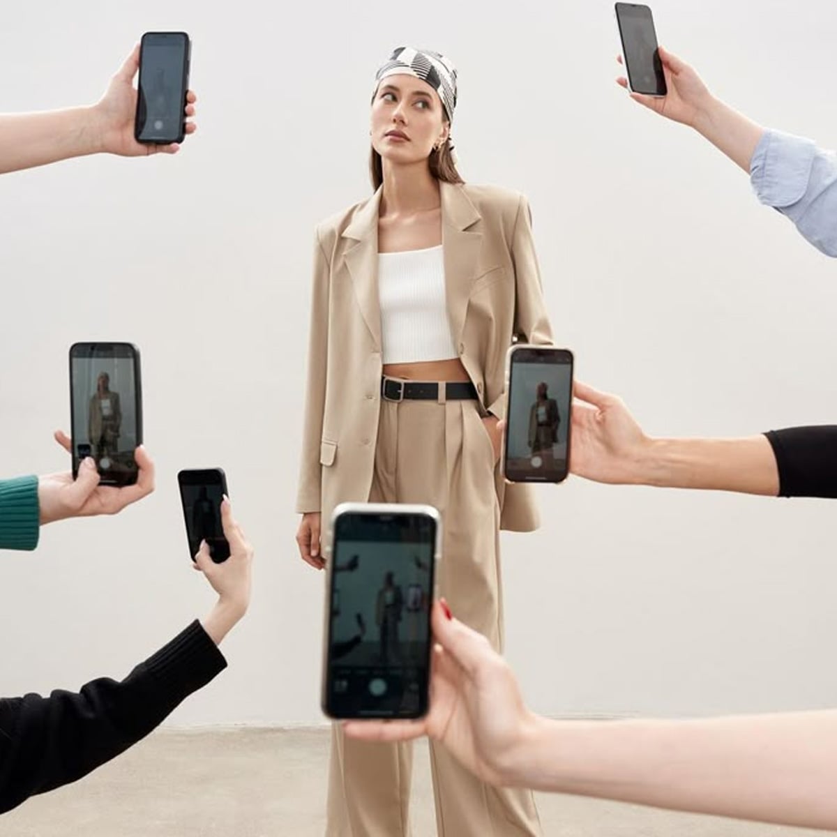 A woman in a beige suit and headscarf stands against a plain background while multiple hands around her take photos of her with smartphones.