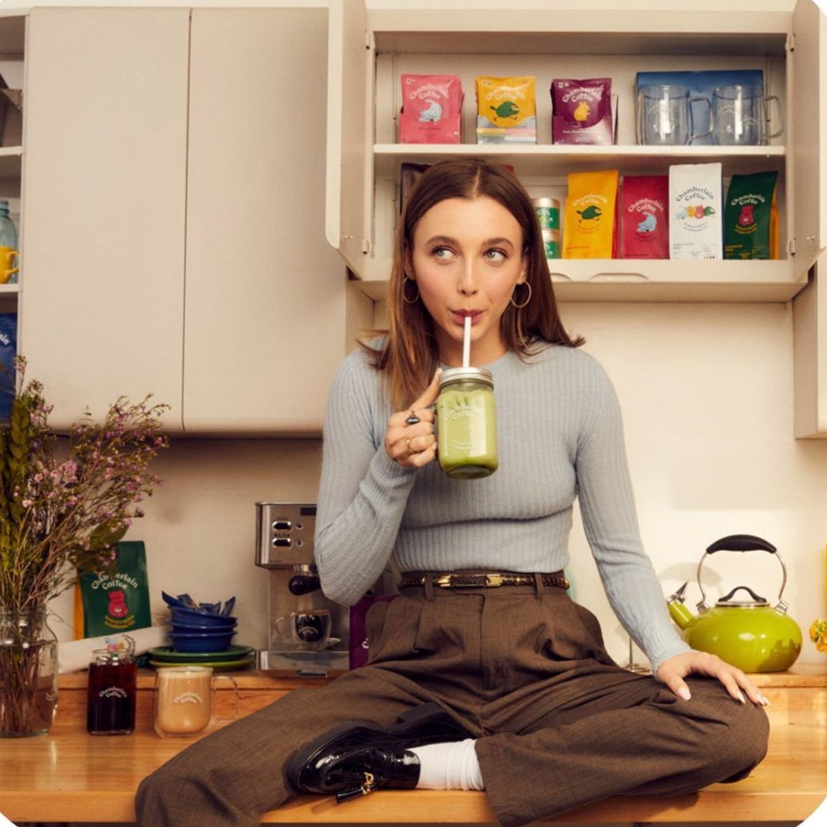A woman sits on a kitchen counter, drinking a green smoothie from a mason jar with a straw. Behind her, shelves display colorful tea packages. She is wearing a light blue sweater and brown pants.