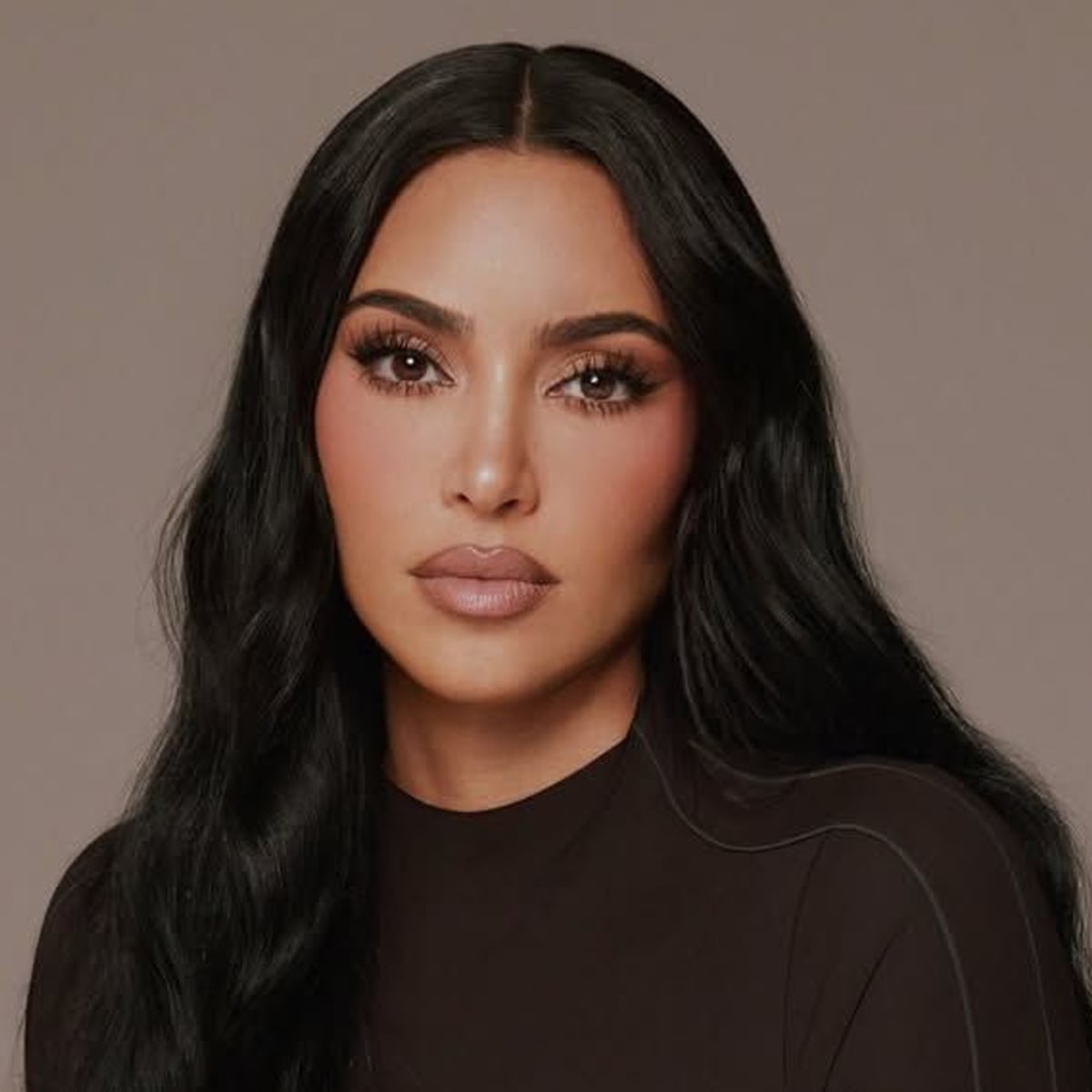A woman with long, wavy black hair and flawless makeup poses against a plain background, wearing a fitted black top and looking directly at the camera with a neutral expression.