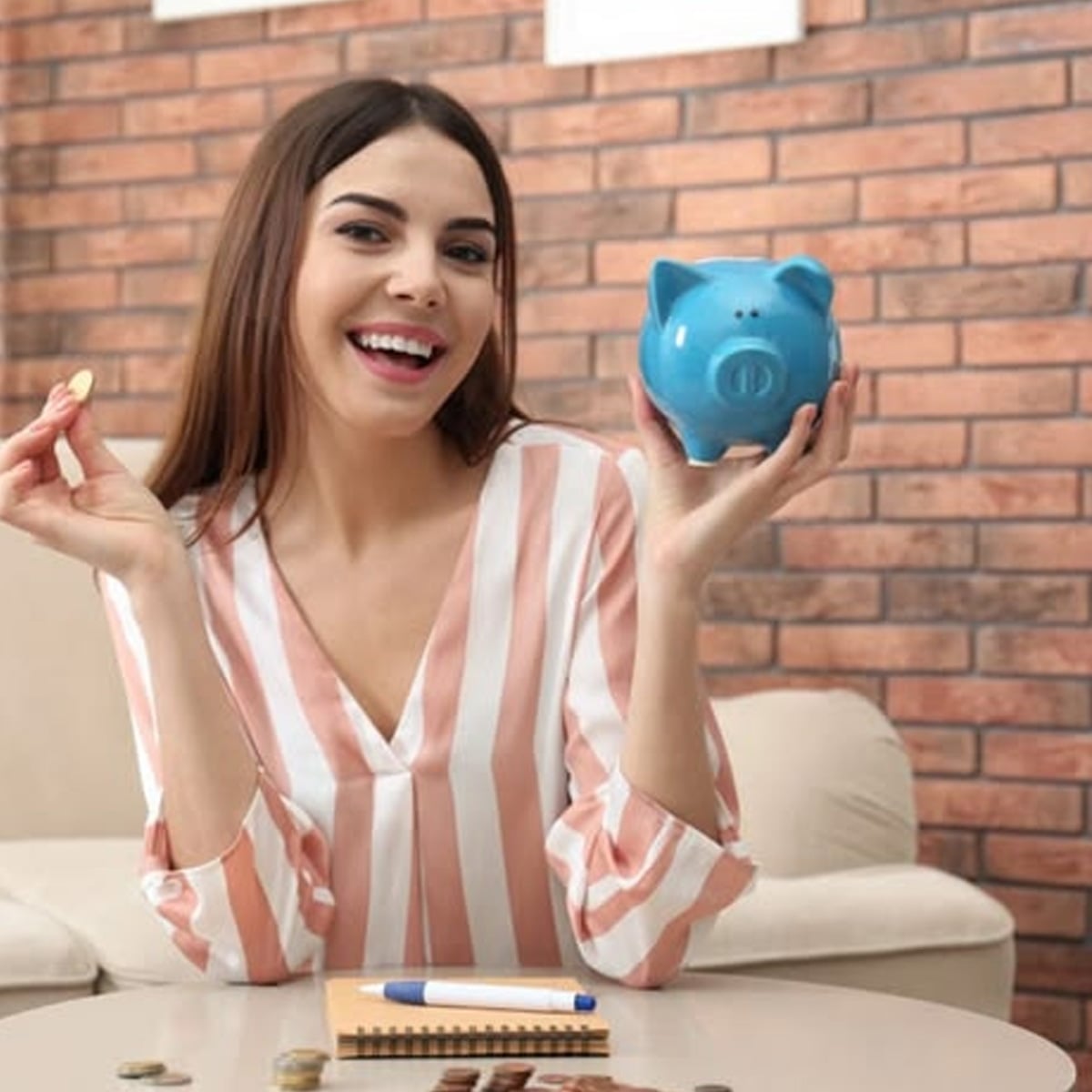 A smiling woman in a striped shirt holds a blue piggy bank in one hand and a coin in the other, sitting at a table with coins, a notebook, and a pen in front of her, against a brick wall background.