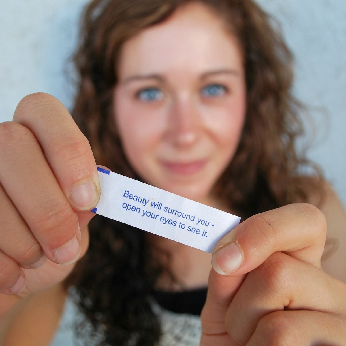 A person with curly brown hair holds a small paper slip close to the camera. The text on the paper reads: "Beauty will surround you - open your eyes to see it." The person smiles softly in the background.