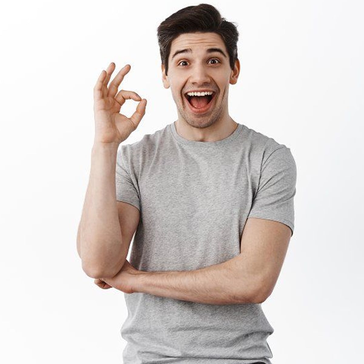A young man with dark hair wearing a grey t-shirt smiles widely and makes an "OK" gesture with his right hand against a plain white background.