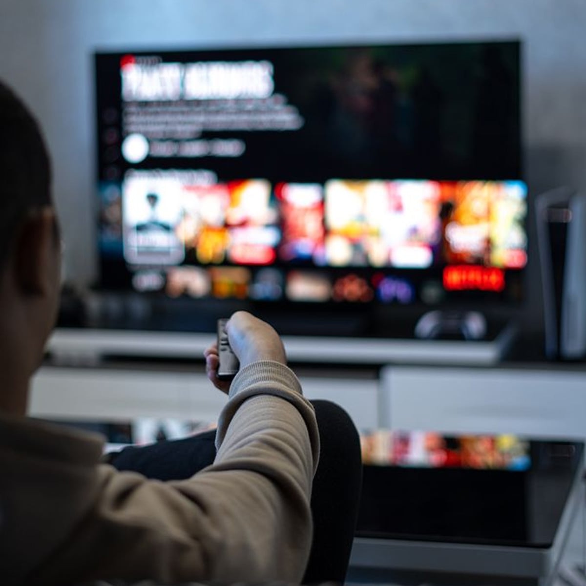 A person sitting on a couch holding a remote control, facing a television displaying a streaming service menu with various movie and show thumbnails.