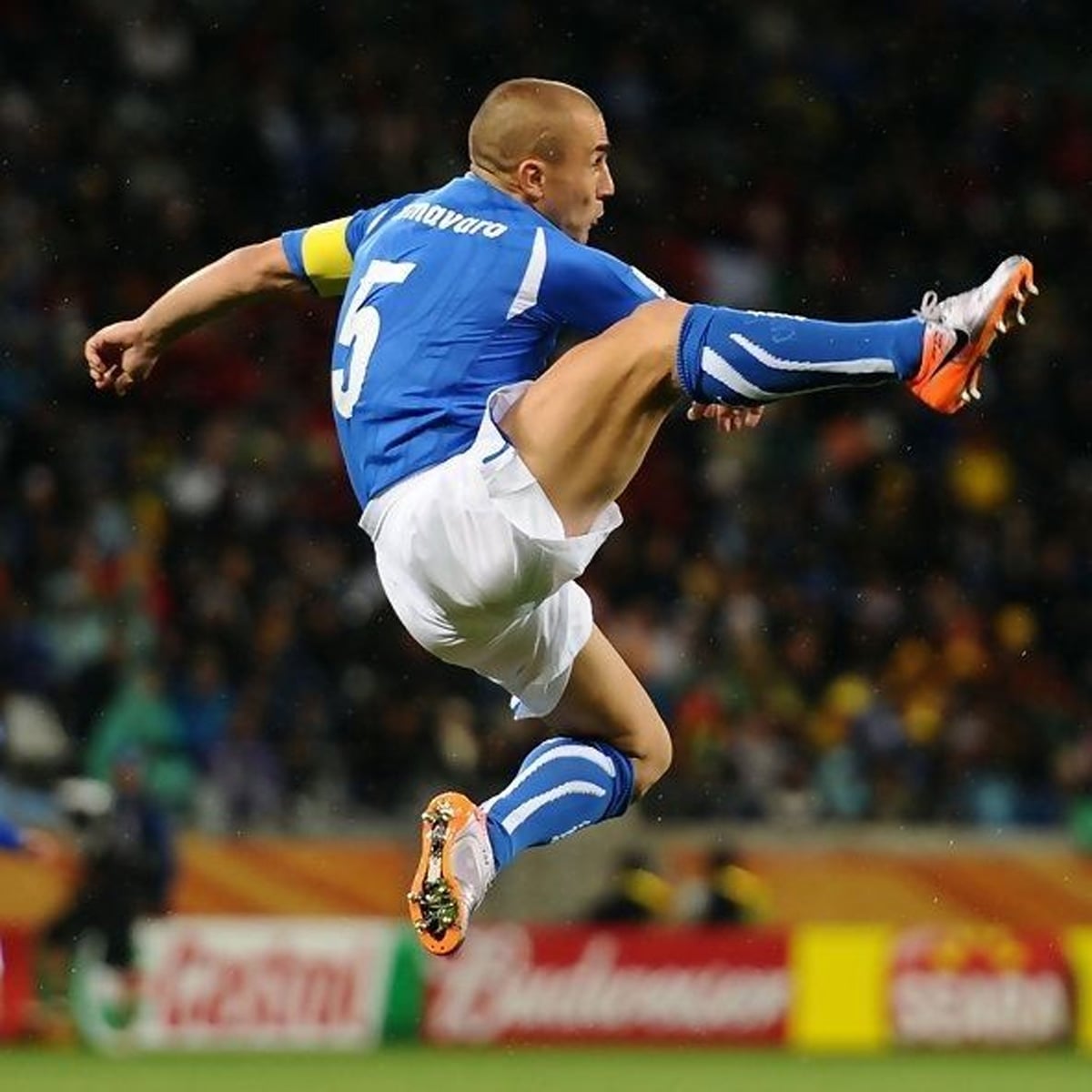 A soccer player in a blue uniform and white shorts leaps into the air, extending his leg for a high kick during a game. The player is wearing a yellow captain’s armband and the number 5 jersey.