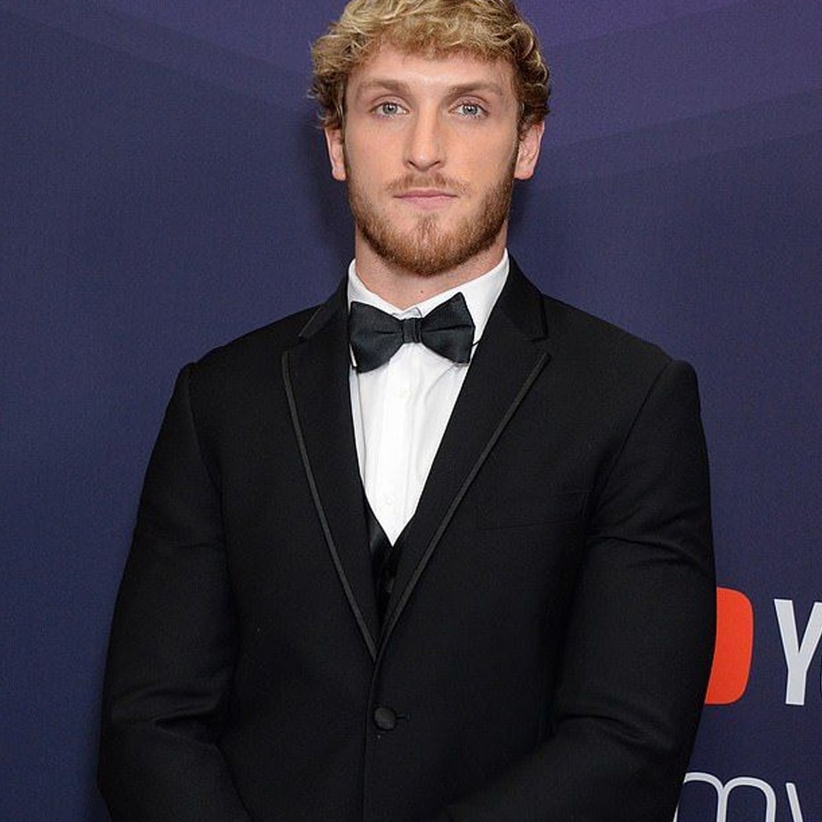 A man with short blond hair and a beard is wearing a black tuxedo and bow tie, standing in front of a purple backdrop with red and white logo elements.