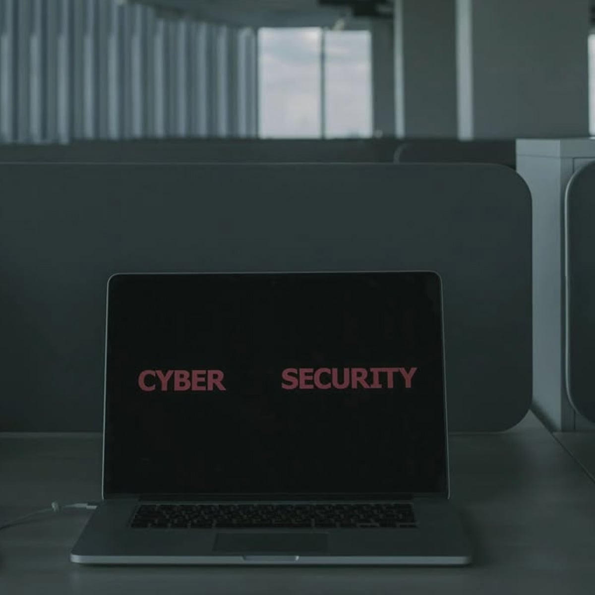 A laptop on a desk in a dimly lit office displays the words "CYBER SECURITY" in bold red letters on its dark screen.