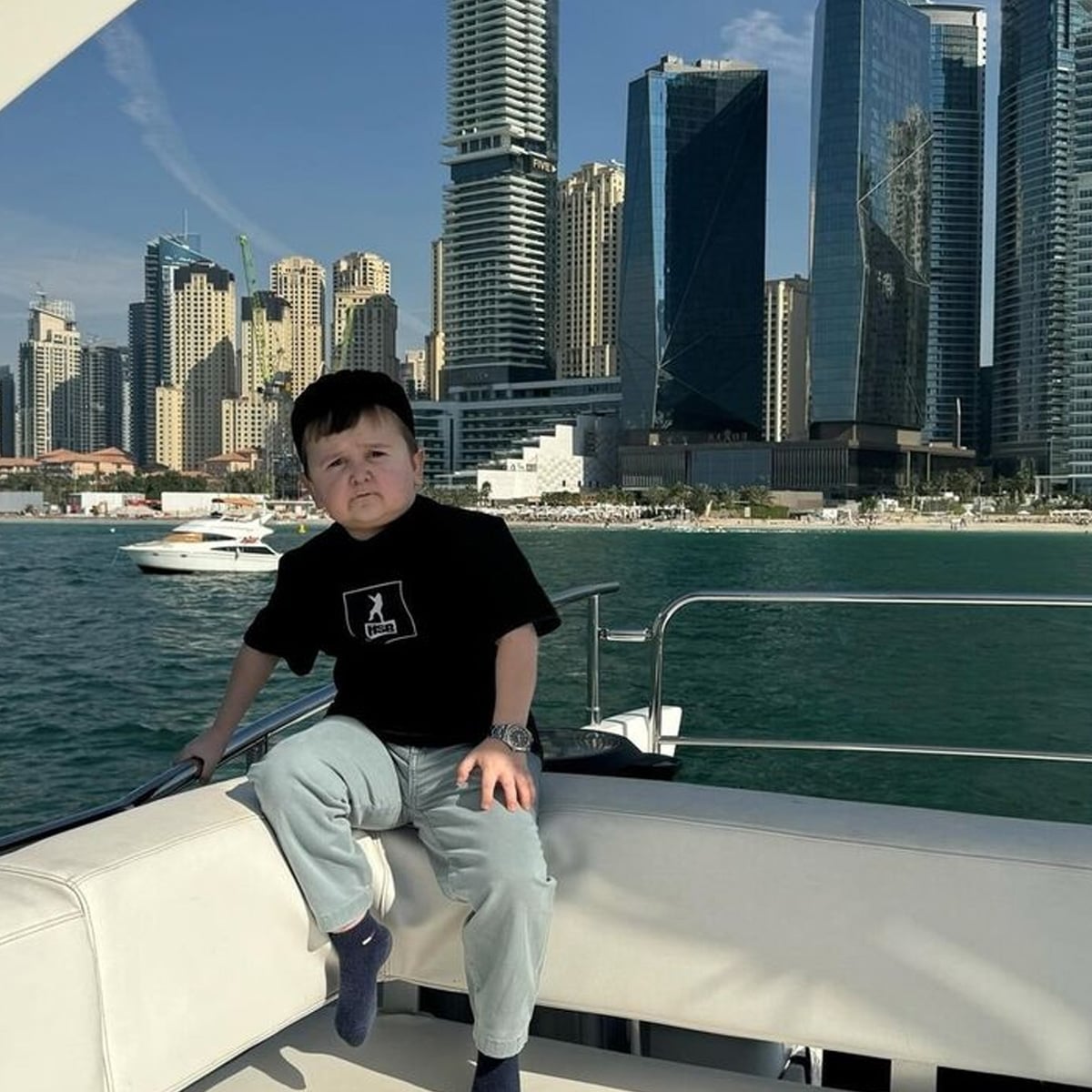 A young boy sits on a yacht with a city skyline of tall, modern buildings in the background. He wears a black t-shirt, light pants, and socks. The yacht is on calm water with another boat nearby.