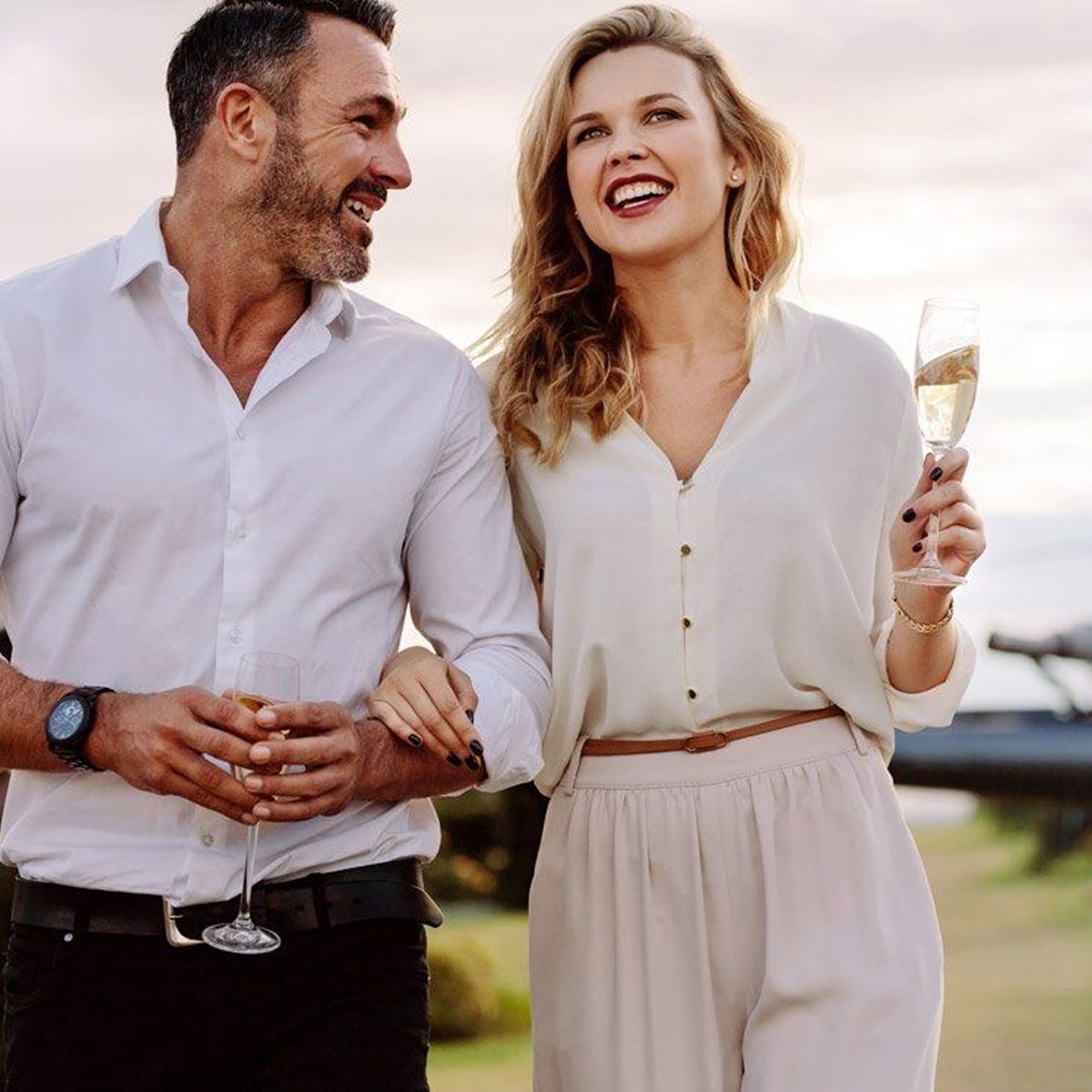 A smiling man and woman dressed in white, holding glasses of champagne, walk outdoors together in the evening light. The woman links her arm through the man's and appears to be laughing.