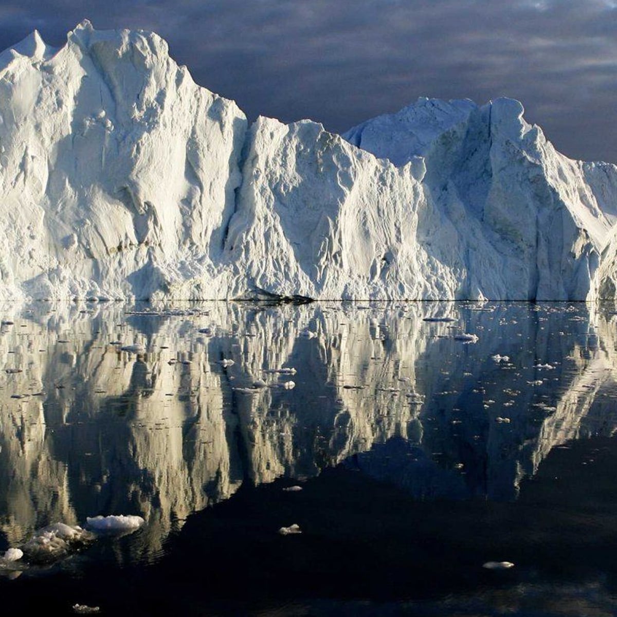 A massive white iceberg rises above dark, calm water, reflecting its jagged, icy surface. Small ice chunks float nearby under a cloudy sky.