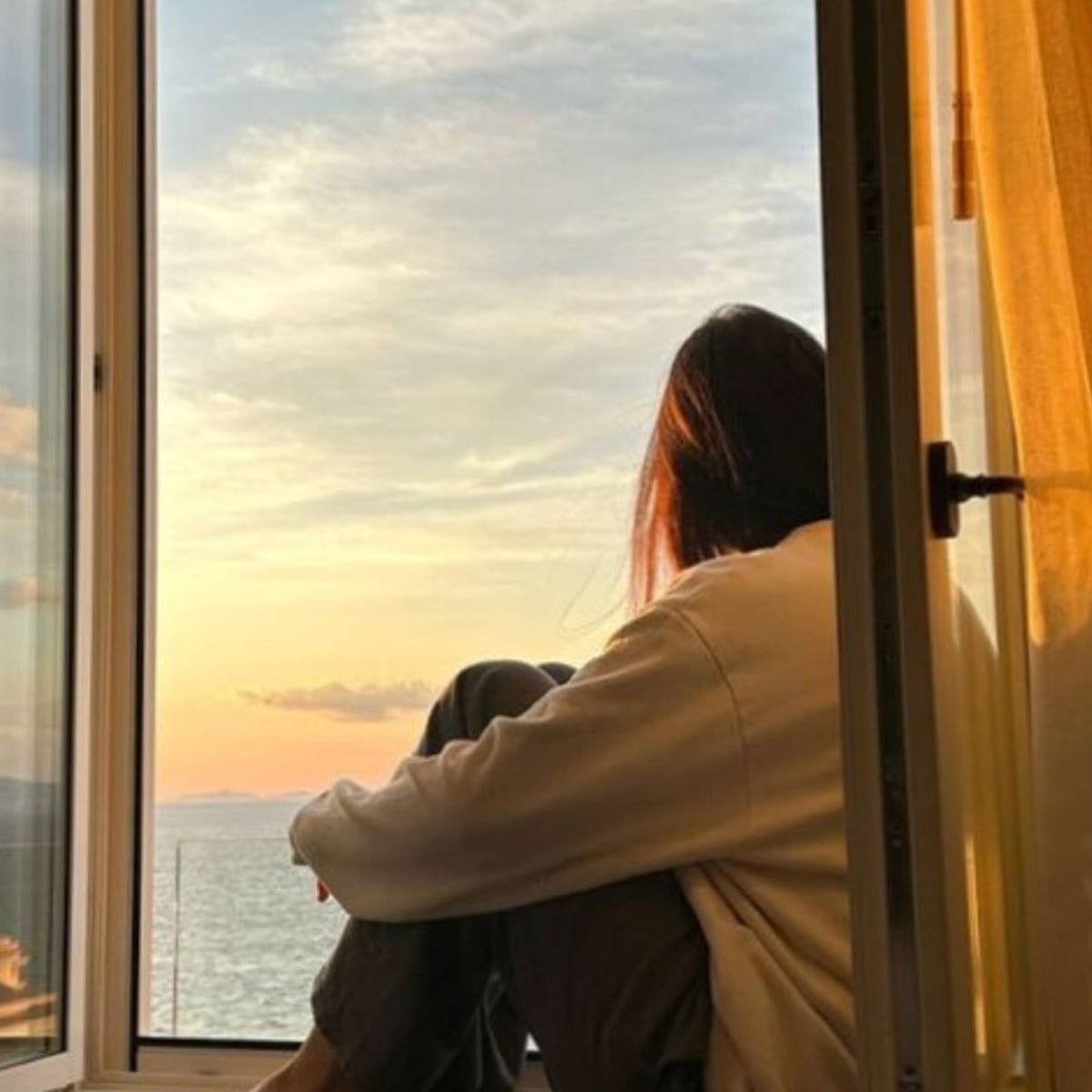 A person sits in a windowsill, gazing outside at a serene sunset over water. Soft golden light fills the room, and the sky is streaked with clouds and warm colors. The person's face is turned away from the camera.
