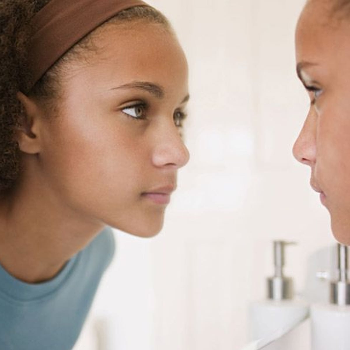 A young person with curly hair and a brown headband looks closely at their reflection in a bathroom mirror, wearing a blue shirt. The background is softly blurred.