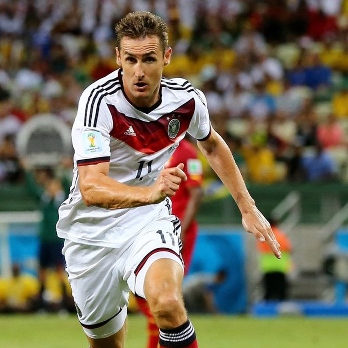A male soccer player in a white and black Germany national team jersey runs on the field during a match, with a blurred crowd and players in the background.