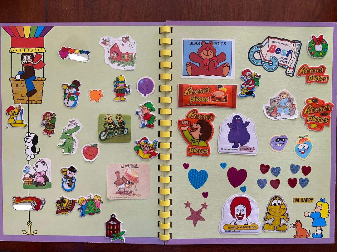 A scrapbook with two pages filled with colorful stickers, including cartoon animals, Sesame Street characters, hearts, stars, Ronald McDonald, Reese’s Pieces, and various cheerful images on a green background.