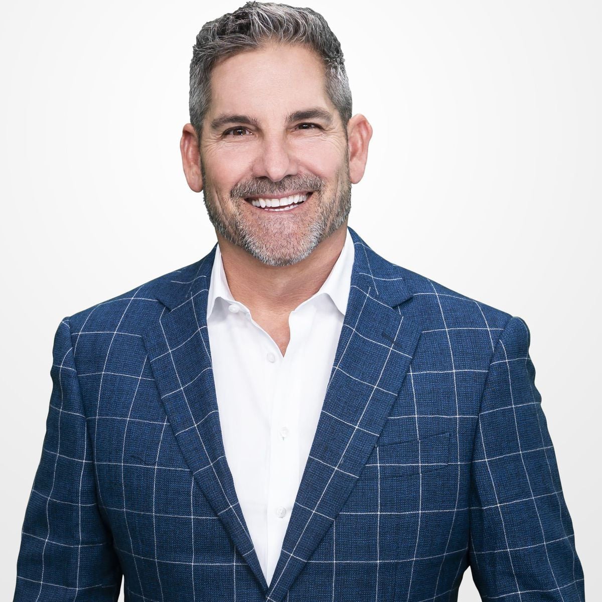 Smiling man with short gray hair and a beard, wearing a blue checkered suit jacket over a white shirt, standing in front of a plain white background.