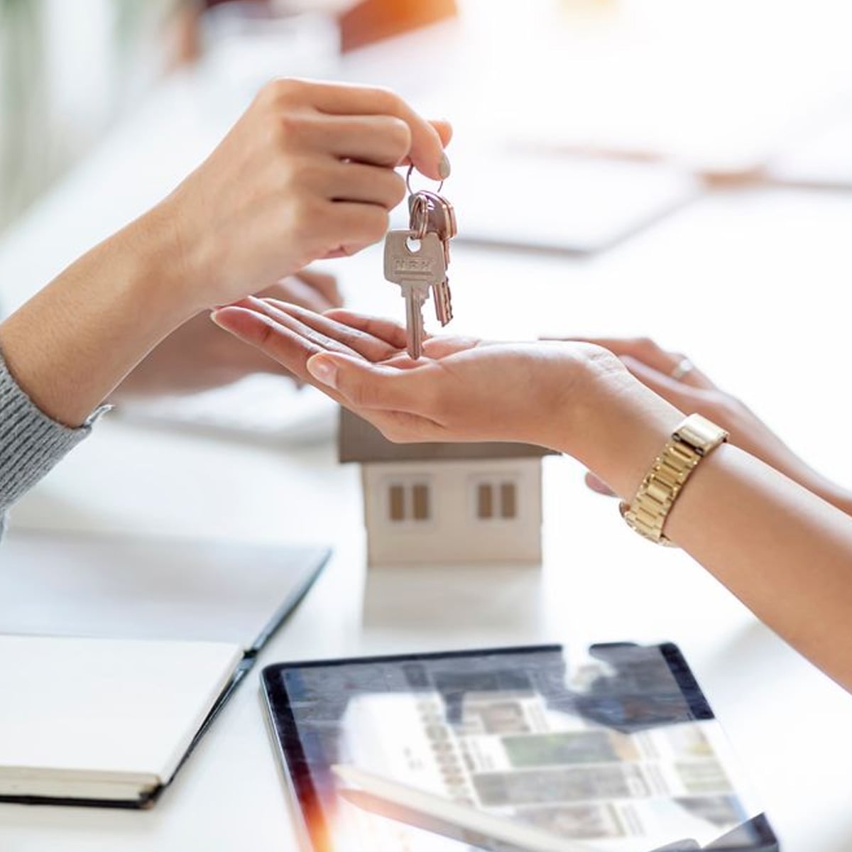 A person hands over a set of keys to another person, symbolizing a property transfer. A small house model, notebook, and tablet are visible on the table in the background.