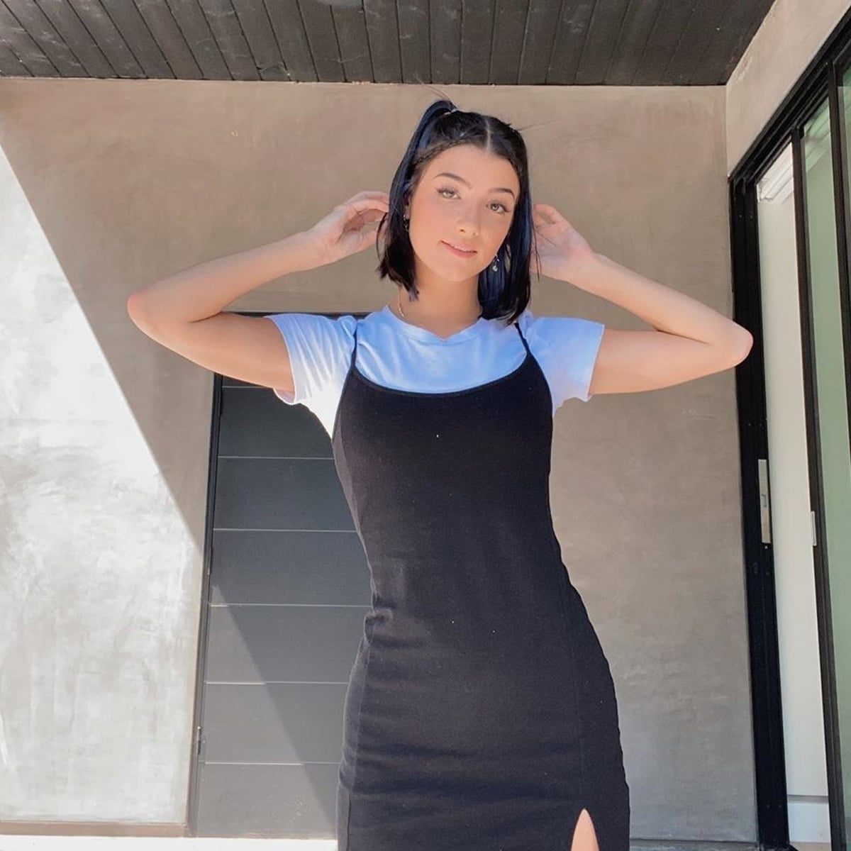 A young woman with dark hair in a half-up hairstyle stands outdoors, posing with her hands near her head. She wears a white t-shirt under a black spaghetti strap dress with a slit. Sunlight creates a shadow on the wall behind her.