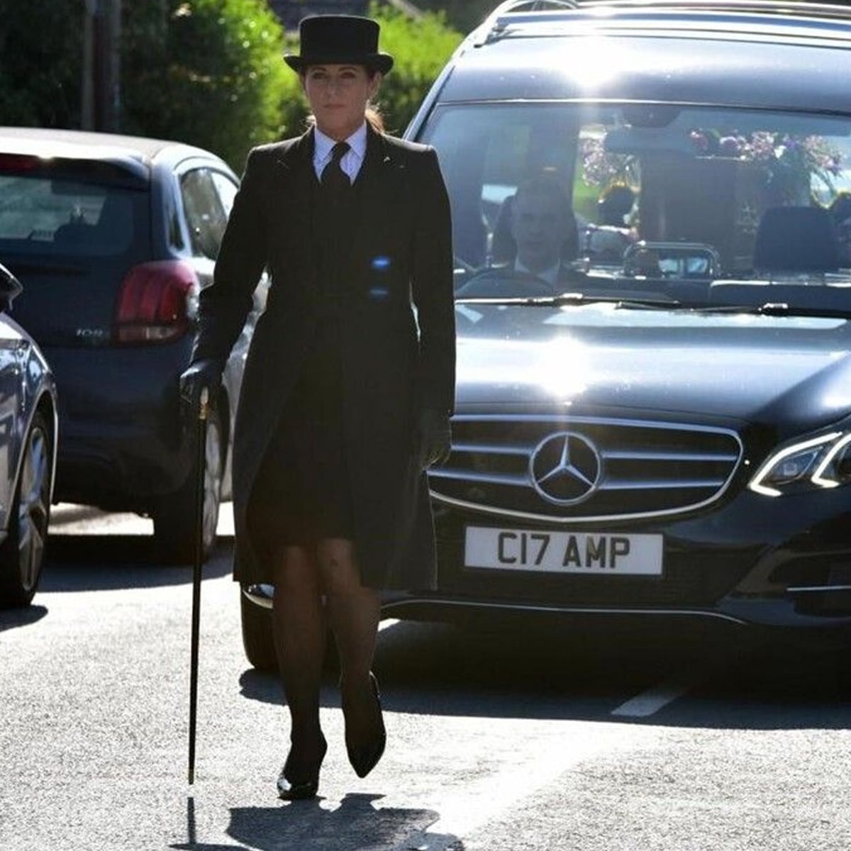 A woman in formal black attire and top hat, holding a cane, walks in front of a Mercedes hearse decorated with flowers, leading a funeral procession on a sunlit street.