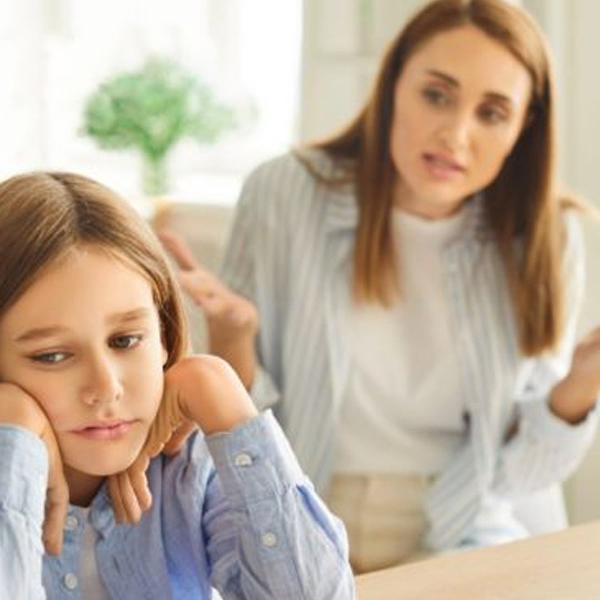 A young girl sits with her chin on her hands, looking upset, while a woman in the background gestures with open hands, appearing concerned or frustrated during a conversation.