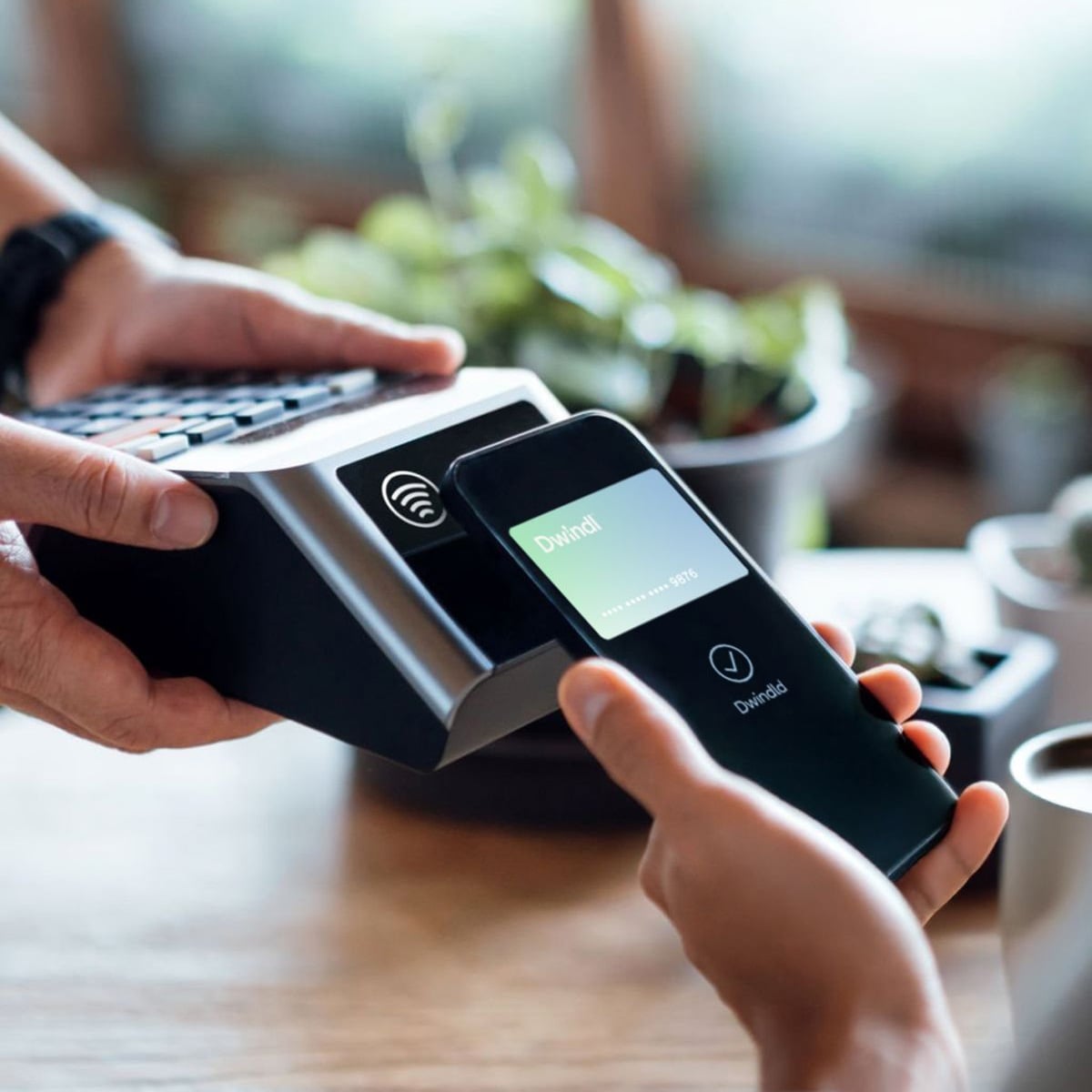 A person holds a smartphone near a card reader to make a contactless payment, with another hand holding the payment terminal; plants are blurred in the background.