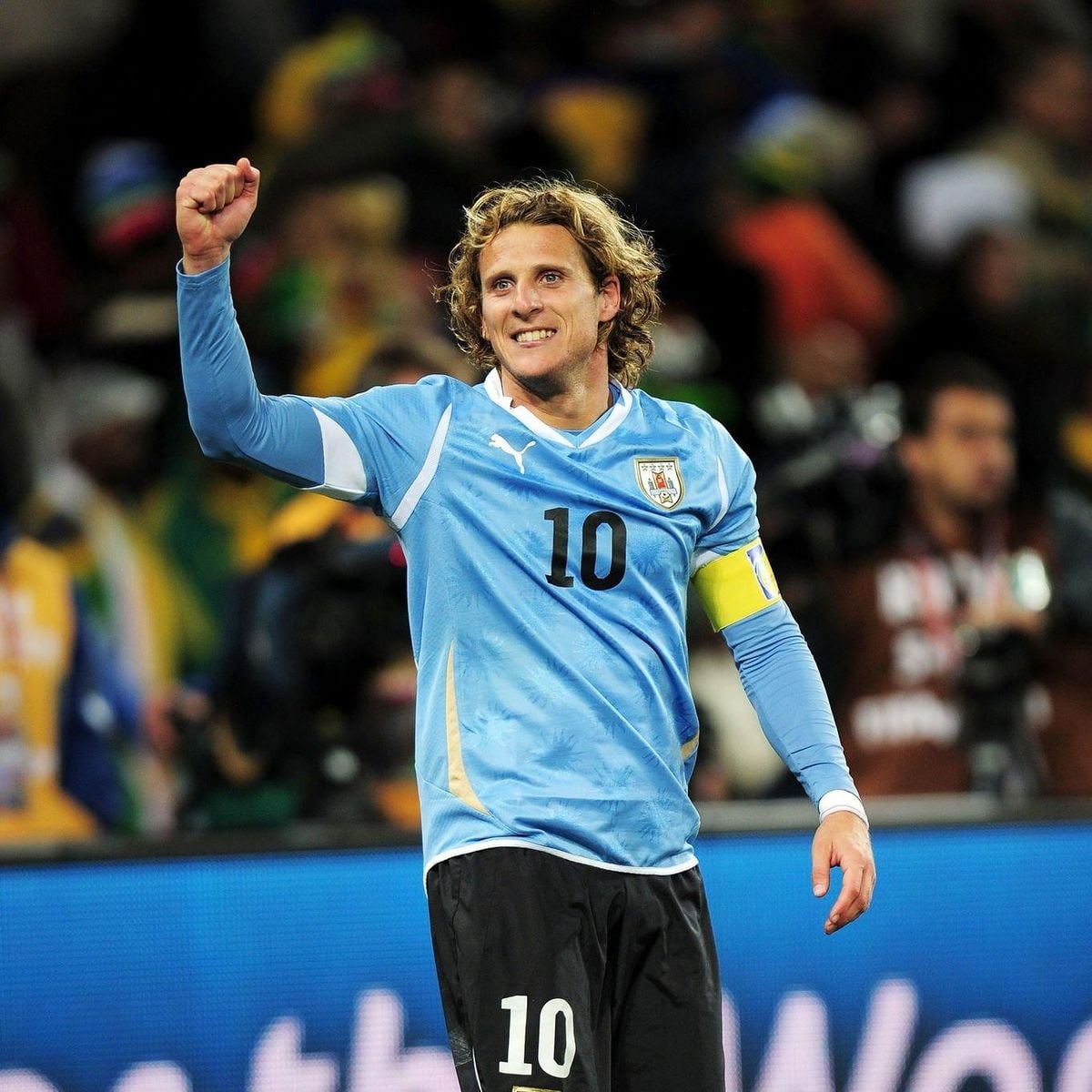 A soccer player in a blue Uruguay jersey with the number 10 celebrates with a raised fist on the field, smiling, with a crowd and cameras in the background.