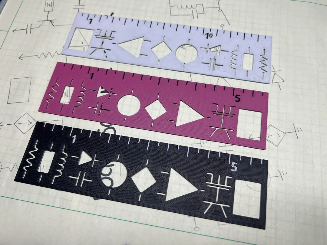 Three colorful circuit diagram stencils—purple, pink, and black—are placed on graph paper featuring hand-drawn electronic symbols. The stencils have cutouts of various electrical components and rulers along the edges.