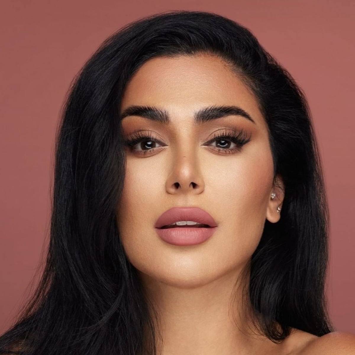 A woman with long black hair, full eyebrows, dramatic eye makeup, and matte pink lipstick poses in front of a solid mauve background.