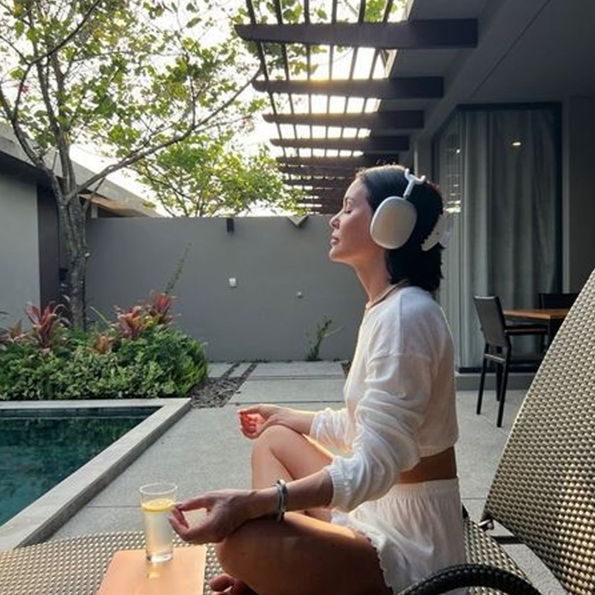 A woman in white loungewear sits cross-legged on a recliner by a pool, wearing large white headphones and holding a drink, with greenery and modern outdoor patio in the background.