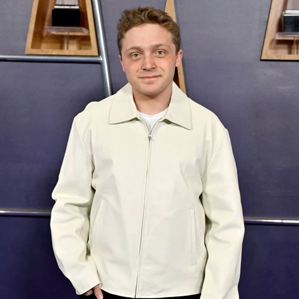 A person with short light brown hair stands in front of a dark wall, wearing a light-colored zip-up jacket. They are looking at the camera with a neutral expression, hands in pockets. Shelves with objects are visible in the background.