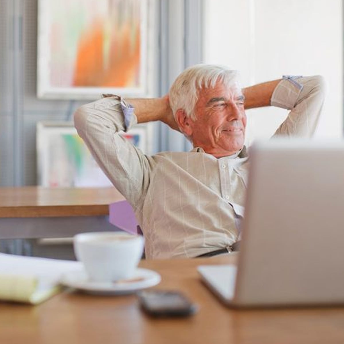 An older man with gray hair sits at a desk with a laptop, coffee cup, notepad, and phone, leaning back in his chair and smiling, looking relaxed in a brightly lit room with abstract art on the walls.