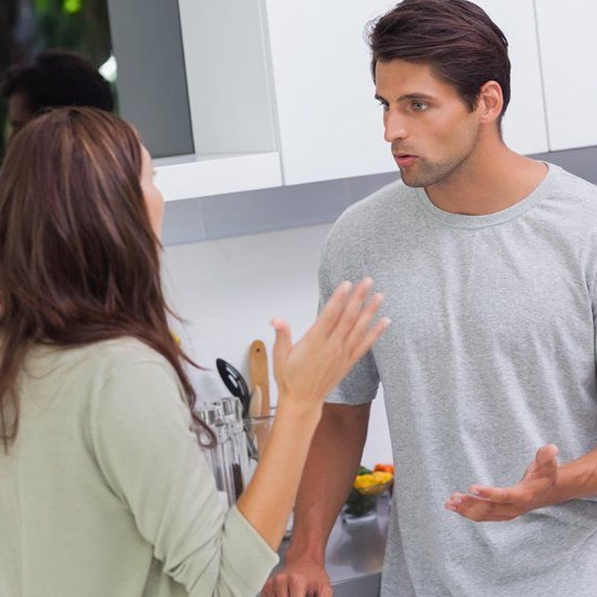 A man and a woman are having a serious conversation in a modern kitchen. The man, wearing a gray t-shirt, gestures with his hand, while the woman raises her hand as she speaks. Both appear engaged in the discussion.