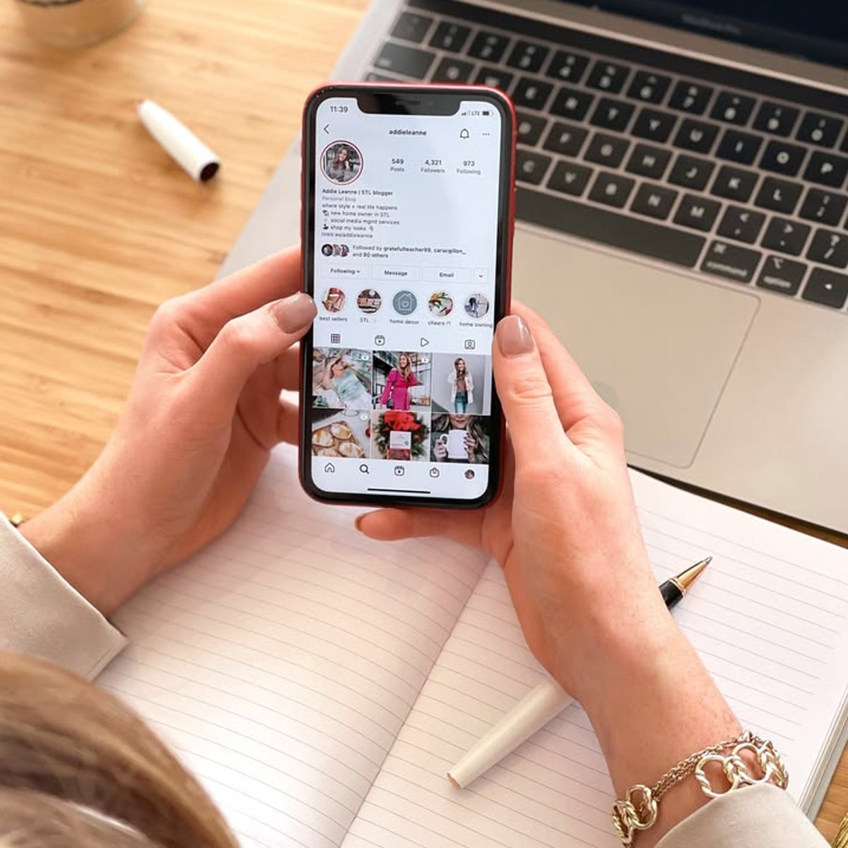 A person holds a smartphone displaying an Instagram profile while sitting at a desk with an open notebook, a pen, and a laptop.