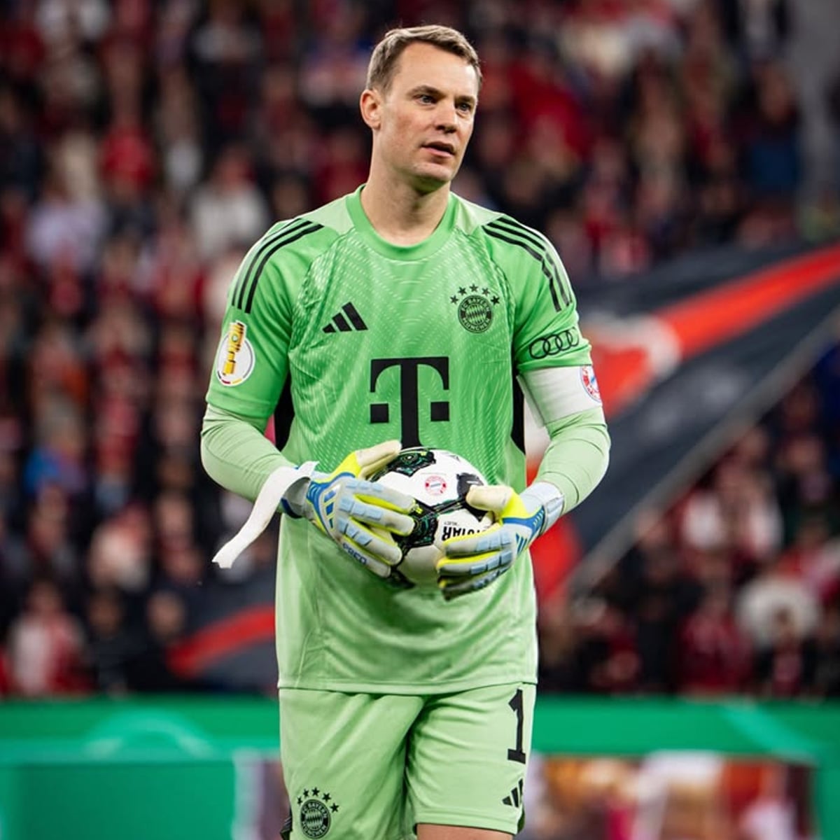 A soccer goalkeeper in a green uniform and gloves holds a ball during a game, with a blurred crowd in the background. The uniform has black stripes and sponsors' logos.