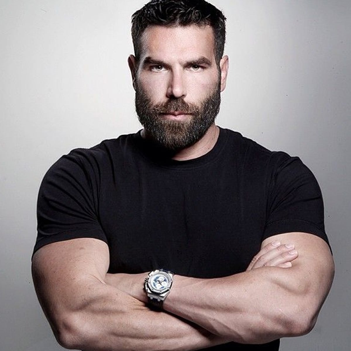 A muscular man with a full beard and short hair stands with his arms crossed, wearing a fitted black t-shirt and a large wristwatch, looking directly at the camera against a plain background.