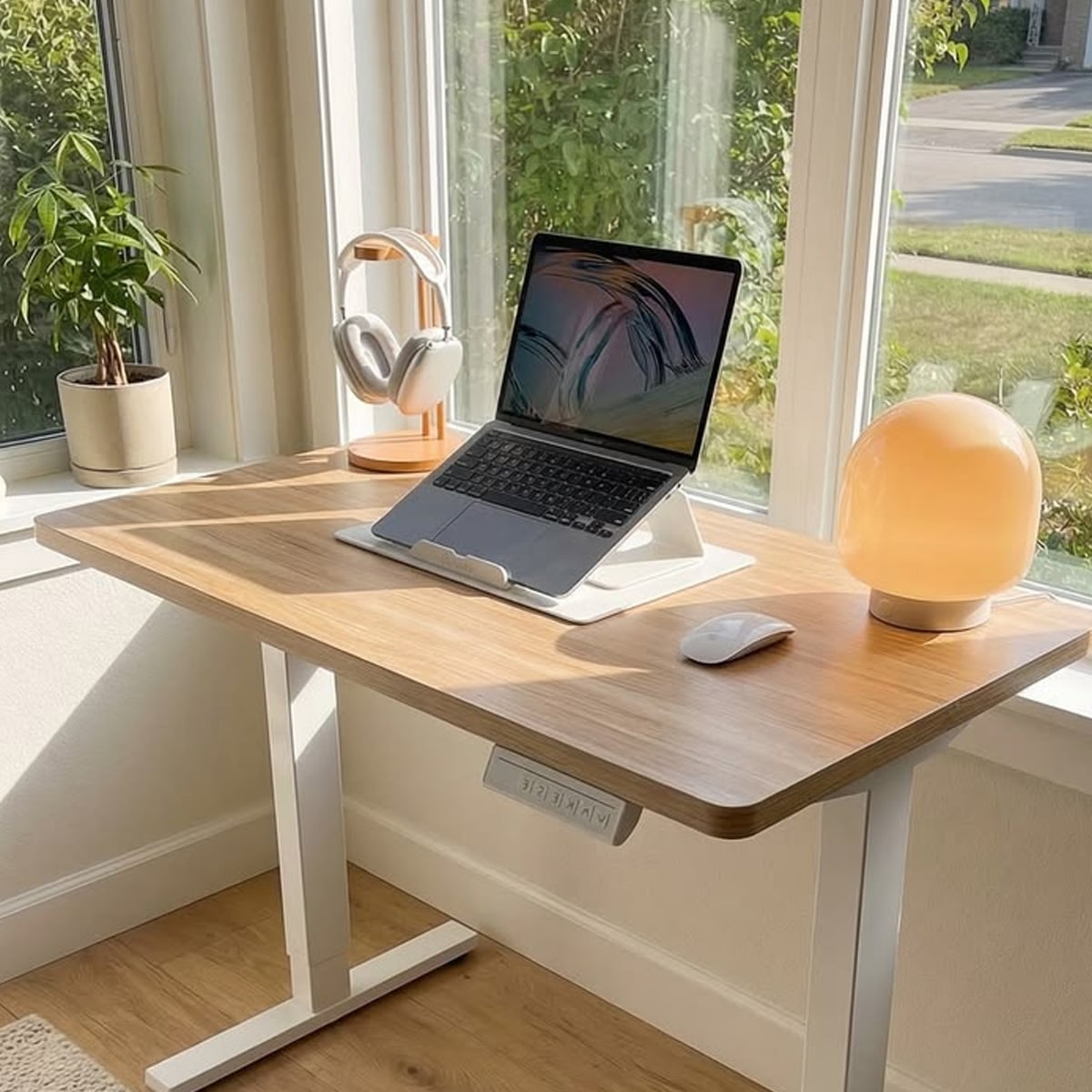 A modern wooden desk by a window holds a laptop on a stand, wireless headphones on a holder, a wireless mouse, a glowing mushroom-shaped lamp, and a small potted plant. Sunlight streams through the windows.