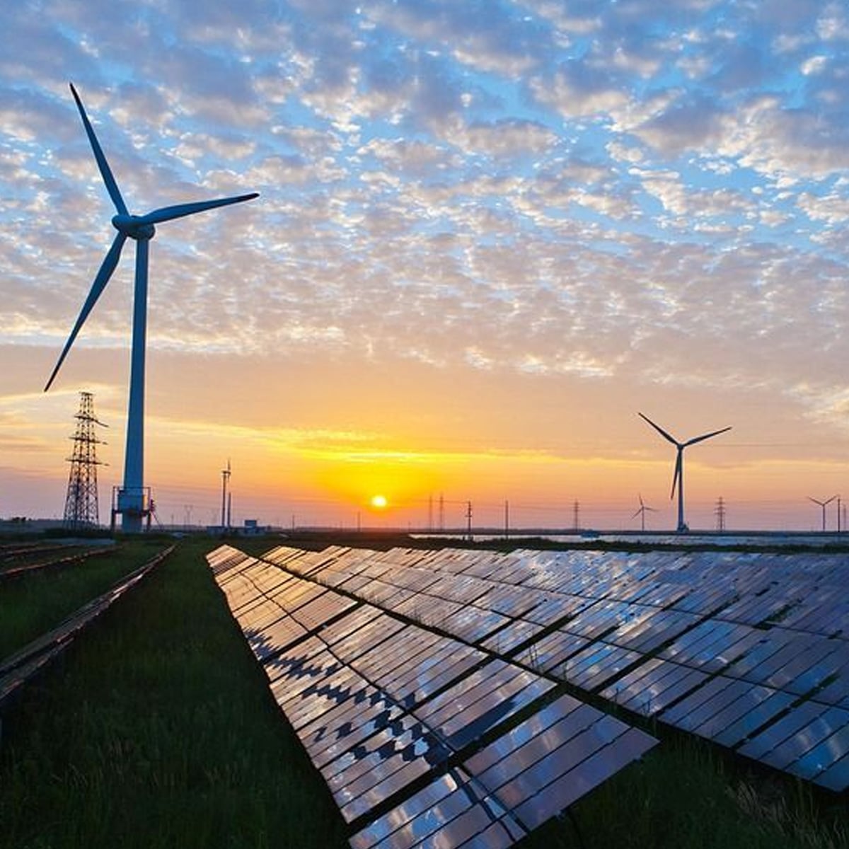 Rows of solar panels and wind turbines stand in a field under a vibrant, cloud-filled sky at sunrise or sunset, representing renewable energy sources.