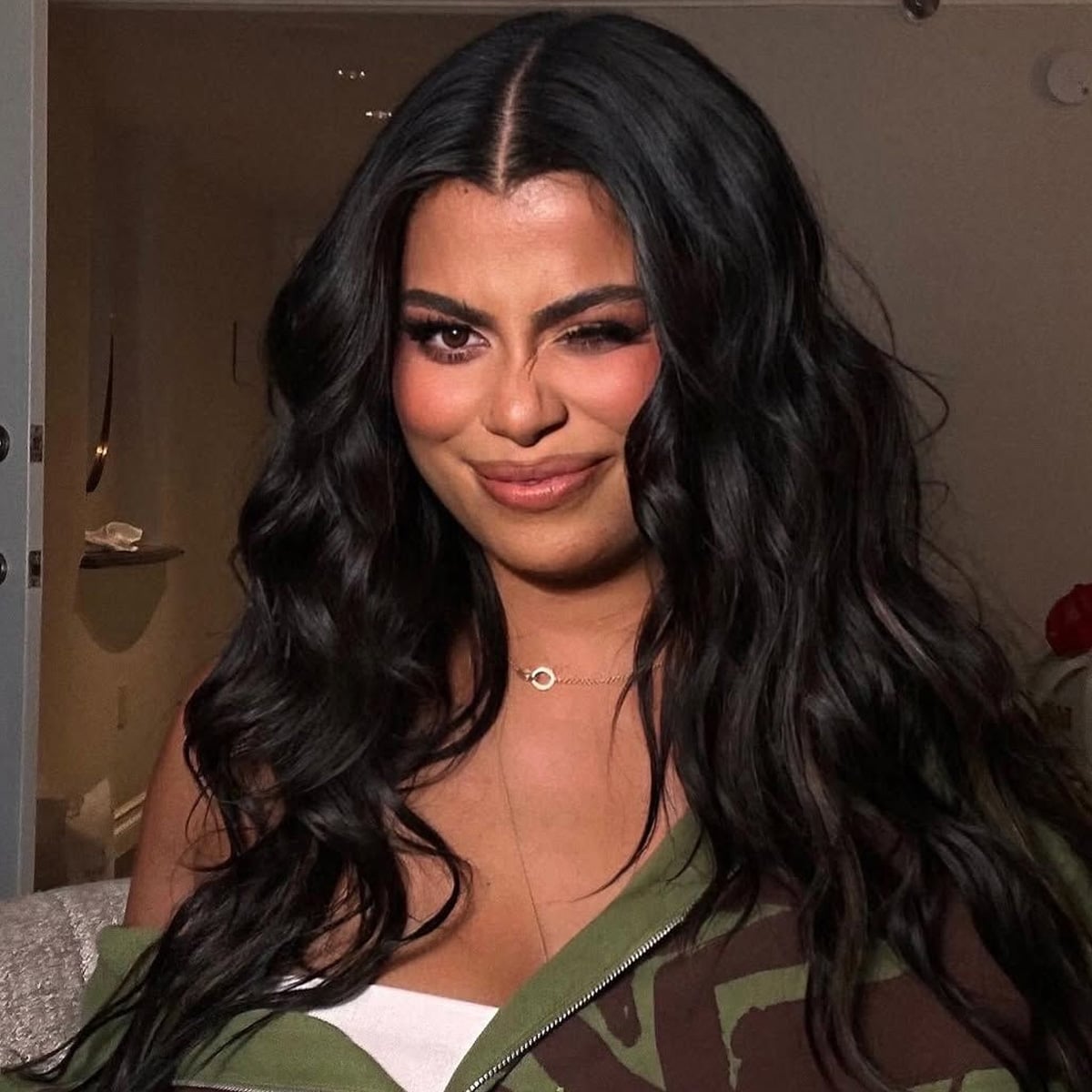 A woman with long, wavy dark hair is indoors, winking and smiling at the camera. She is wearing a green jacket over a white top and a delicate necklace. The background shows a dimly lit room.