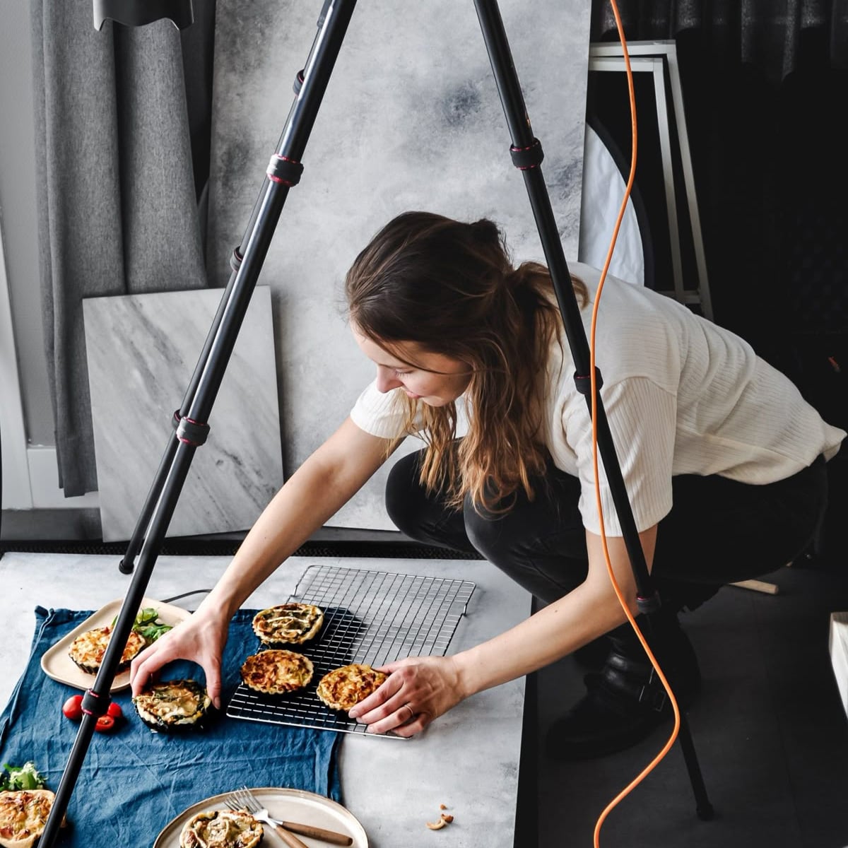A woman arranges food on a cooling rack for a photo shoot, with a camera tripod and cord in the foreground. Plates of food and a blue cloth are spread on the floor around her.