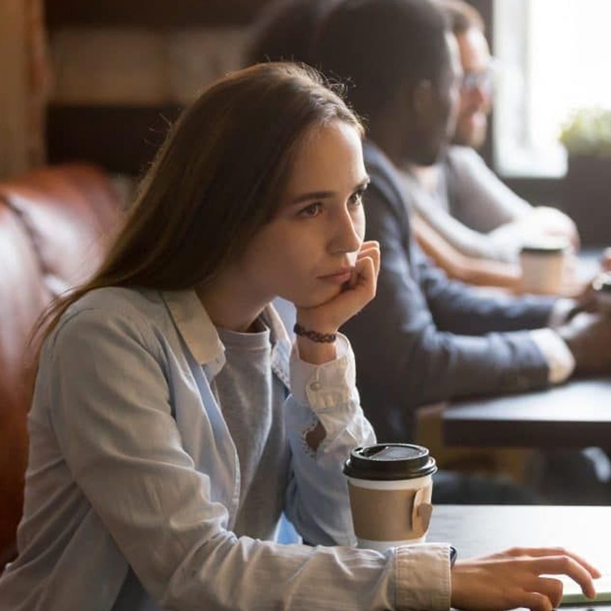 A young woman sits at a table in a café, resting her chin on her hand and looking thoughtful. She has long brown hair and is wearing a light blue shirt. A takeaway coffee cup is on the table in front of her.