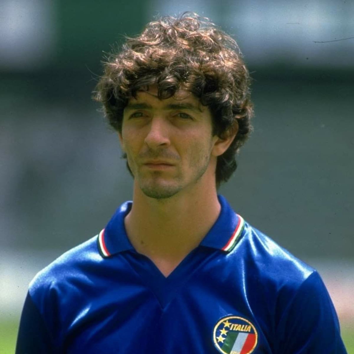 A male soccer player with curly brown hair wears a blue Italy national team jersey with the Italian flag emblem, standing outdoors with a neutral expression.