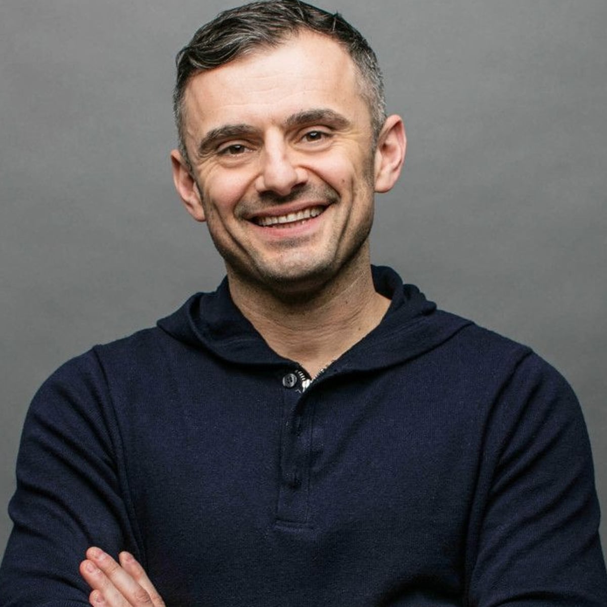 A man with short dark hair and a light beard is smiling with his arms crossed, wearing a dark blue henley shirt against a plain gray background.