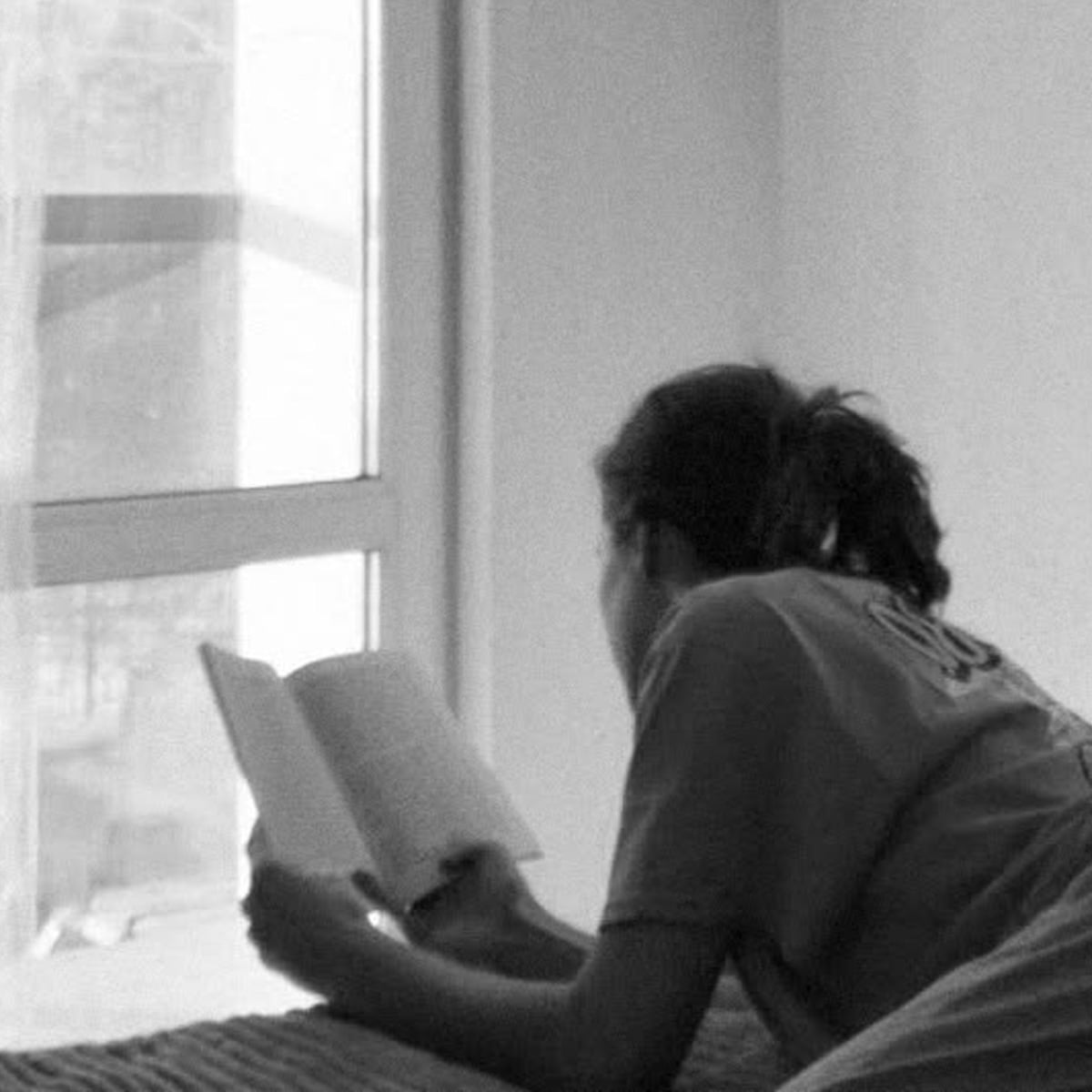 A person lies on their stomach on a bed near a window, reading a book. The scene is in grayscale, and natural light comes through the window, creating a calm and relaxed atmosphere.