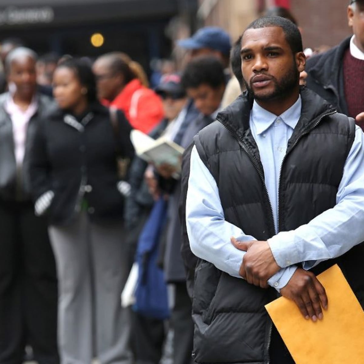 A man holding a large brown envelope stands at the front of a line of people outdoors. The group, dressed in business attire, appears to be waiting for something, with some reading or looking ahead.