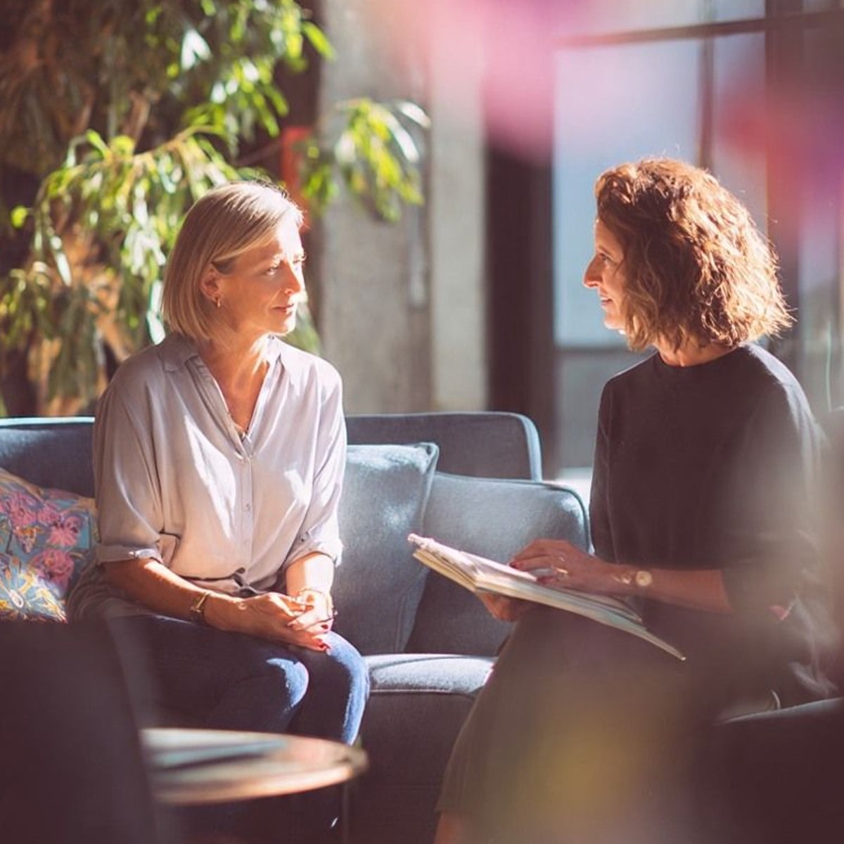 Two women sit on a blue sofa in a sunlit room, engaged in conversation. One woman holds an open book or folder, and both appear focused and relaxed. Green plants and large windows are visible in the background.