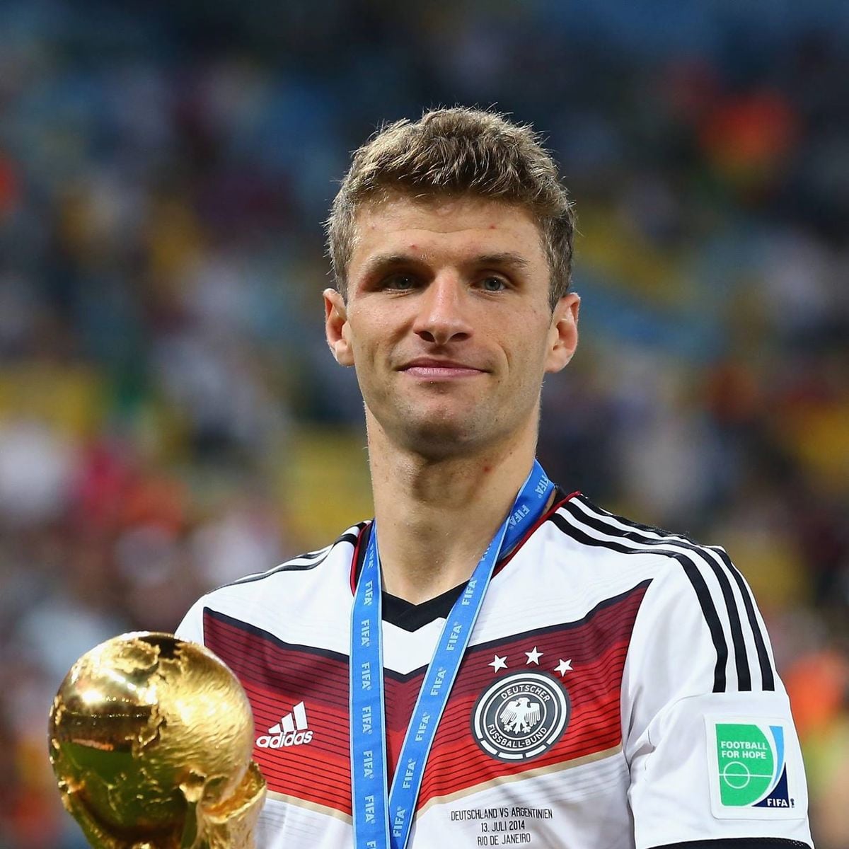 A soccer player in a white Germany jersey with a medal around his neck holds the FIFA World Cup trophy, standing in a stadium with blurred spectators and bright lights in the background.