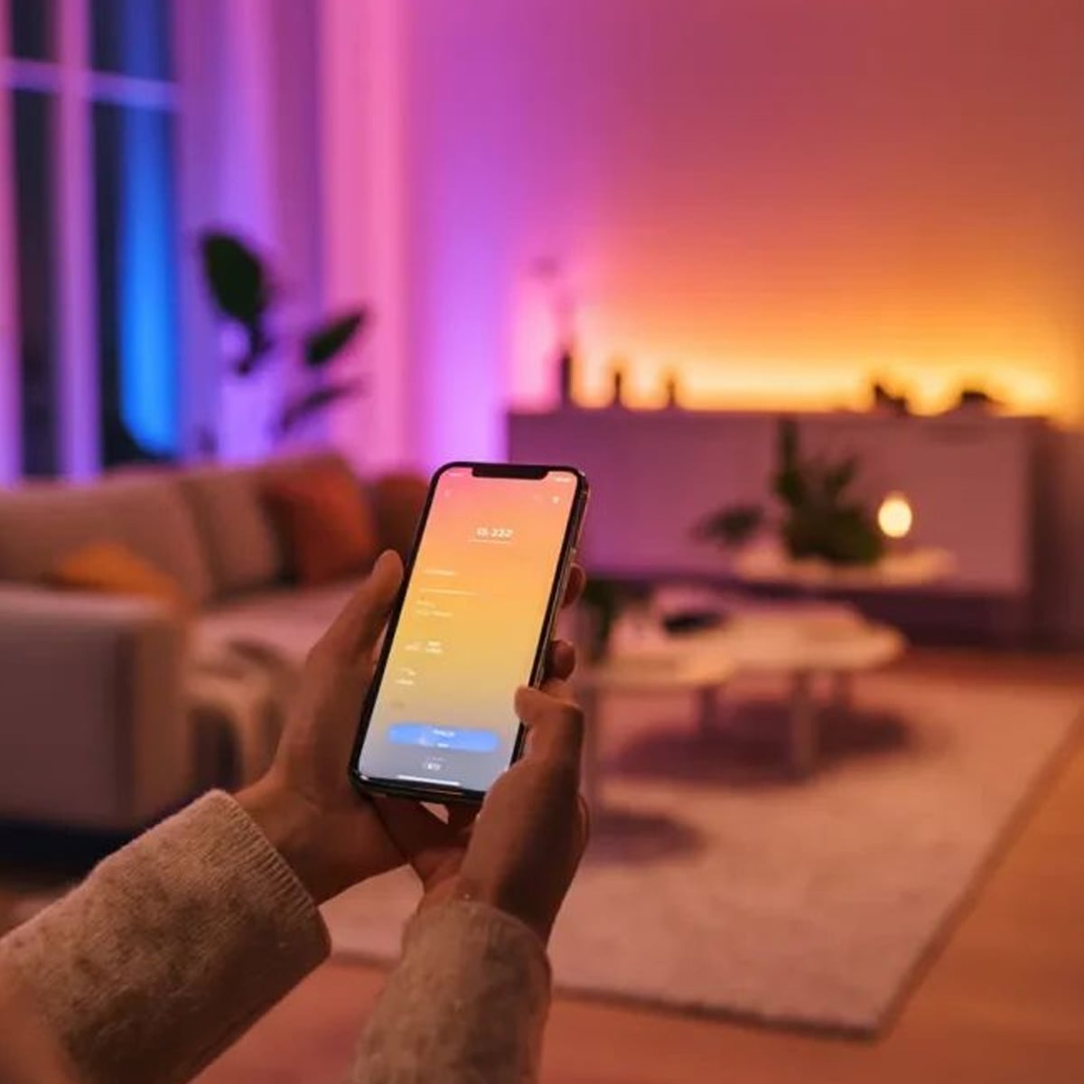 A person holds a smartphone controlling ambient lighting in a cozy living room with colorful, soft purple and orange lights, modern furniture, and plants visible in the background.