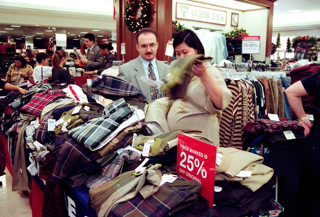 Shoppers browse through piles of discounted clothes in a busy department store decorated for the holidays, with a sign showing a 25% off sale in the foreground.