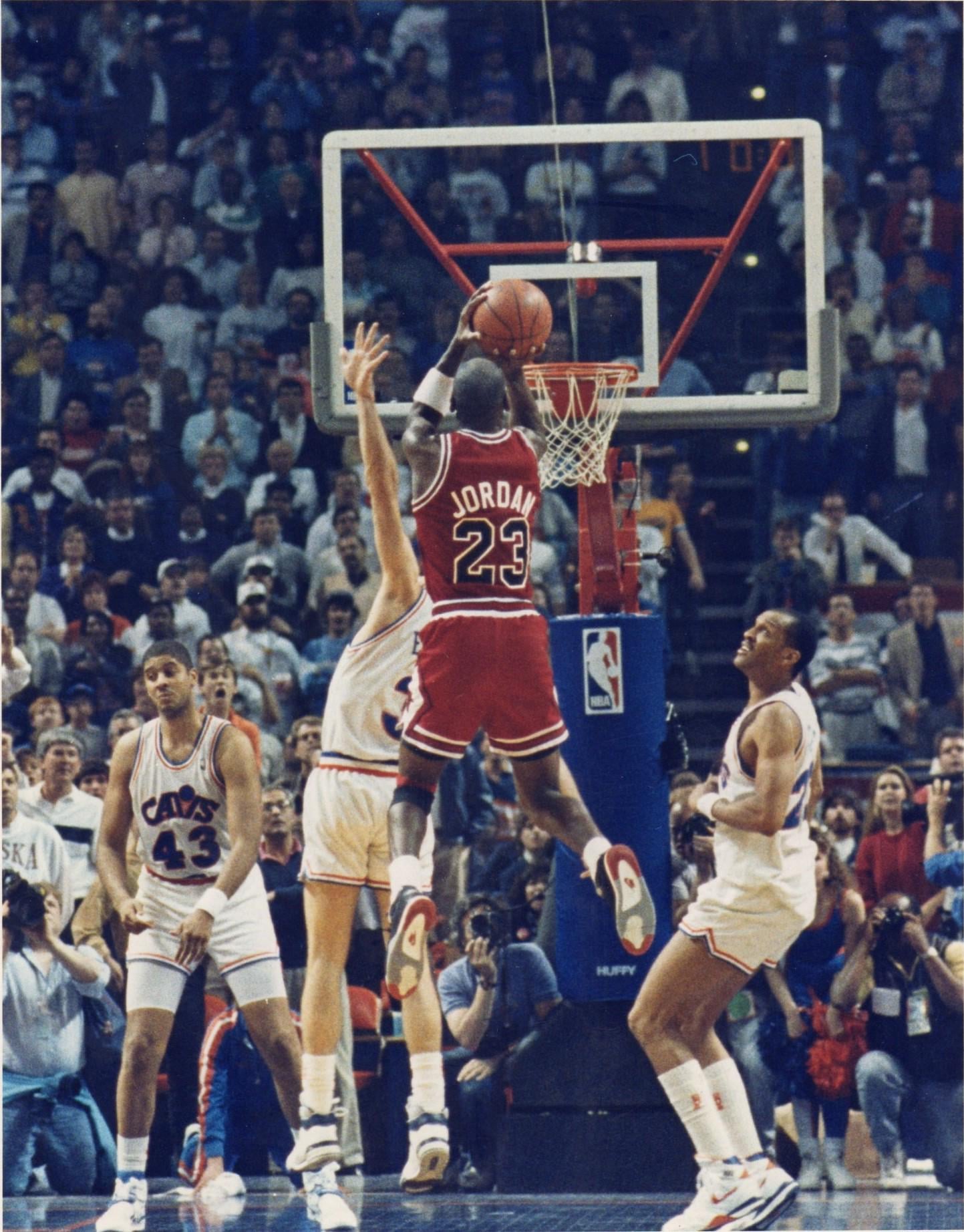 A basketball player in a red jersey, number 23, jumps to take a shot while being closely defended by a player in a white jersey during a crowded game, with spectators watching intently.