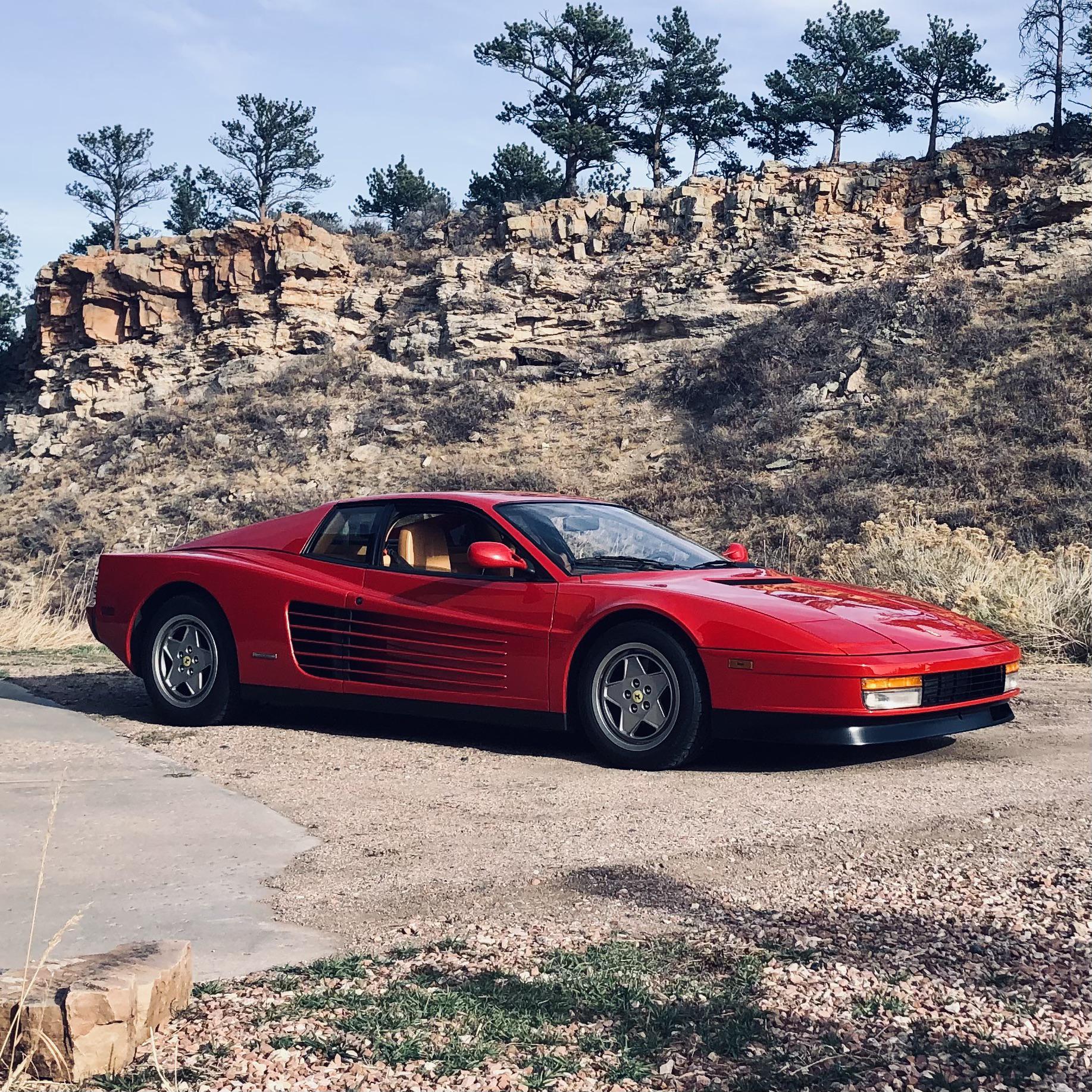 A red Ferrari sports car is parked on a paved area beside a rocky hillside with sparse trees and dry grass under a partly cloudy sky.